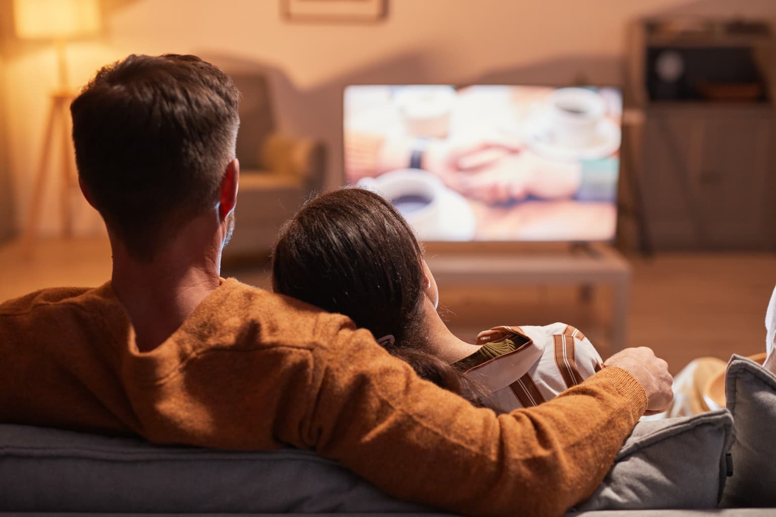 Zwei Personen sitzen auf einem Sofa, mit dem Rücken zur Kamera, und schauen fern. Die linke Person hat den Arm um die rechte gelegt. Auf dem Fernseher ist ein Bild von Händen mit Kaffeetassen zu sehen. Der Raum ist weich beleuchtet, im Hintergrund stehen eine Lampe, ein Tisch und Regale.