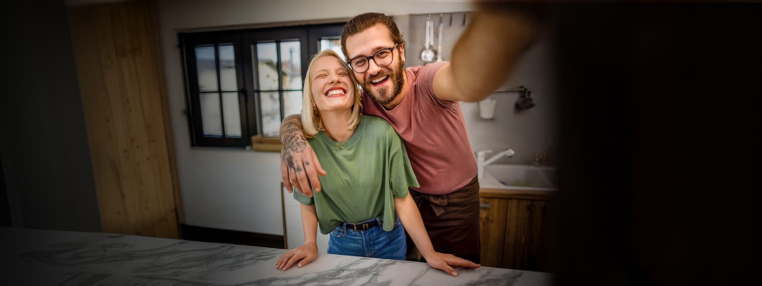 Lächelndes Paar macht Selfie in moderner Küche mit Holzelementen und Marmorarbeitsplatte.