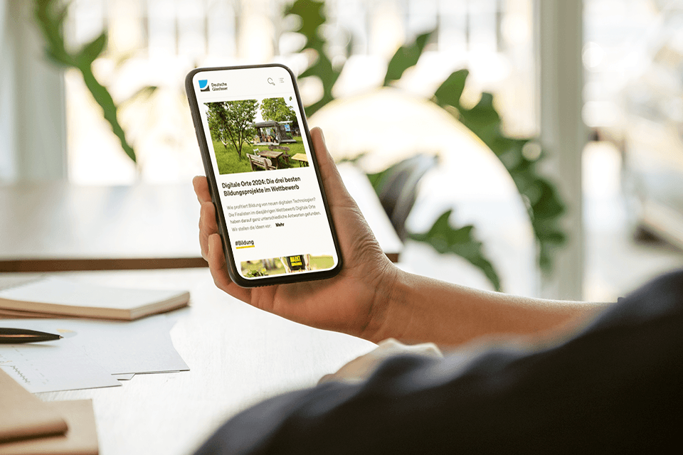 Person hält Handy am Schreibtisch in der Hand und schaut sich die Digitales Bürgernetz Seite an