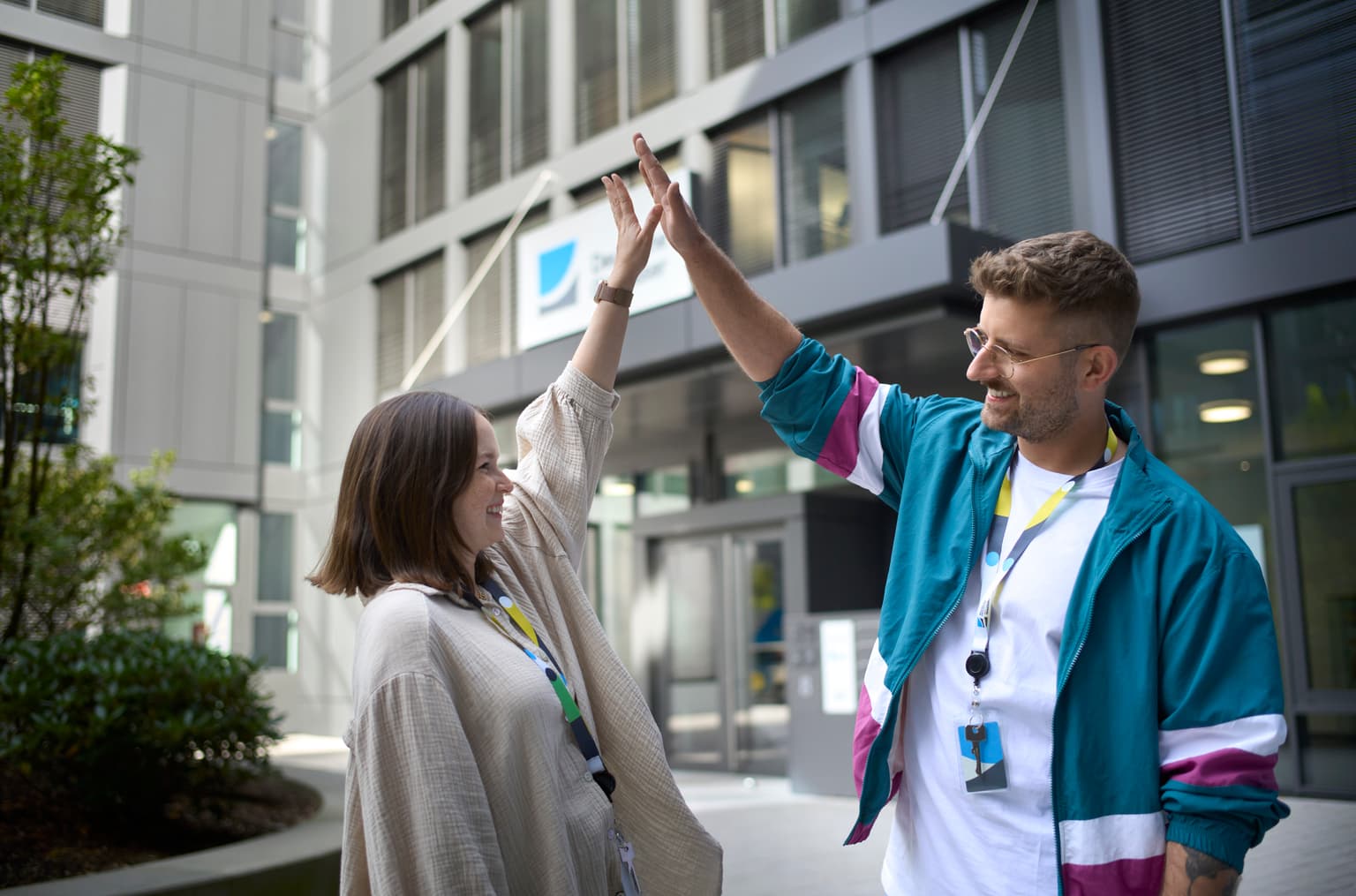 Das Bild zeigt zwei Mitarbeitende, die draußen vor dem Bürogebäude stehen, lachen und sich Highfive geben.
