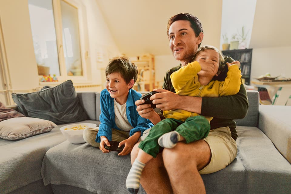 Ein Vater sitzt mit zwei Kindern auf dem Sofa und spielt Videospiele. Alle lachen und halten Controller in den Händen.