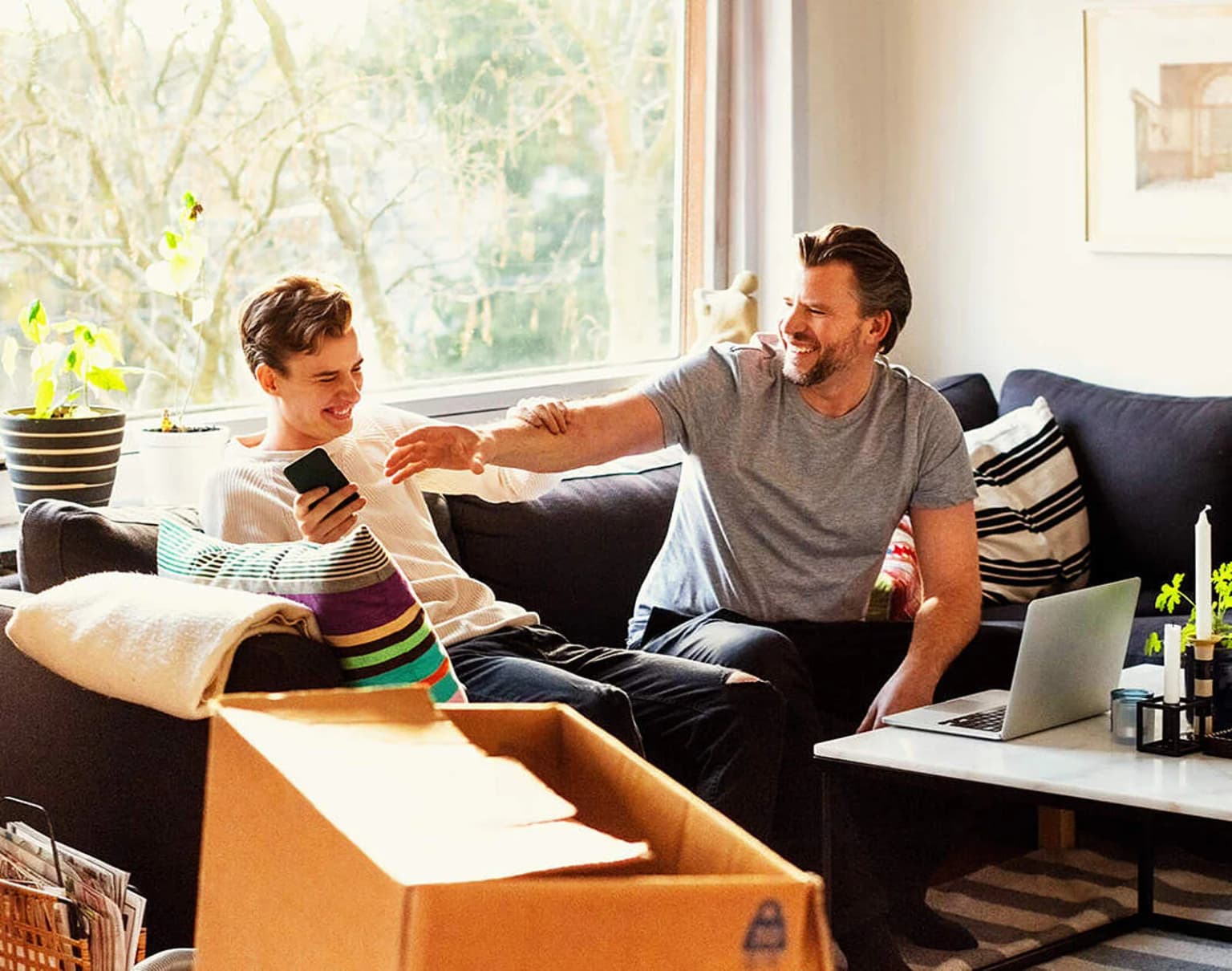 Vater und Sohn sitzen auf der Couch im Wohnzimmer und lachen. Vater möchte seinem Sohn das Handy aus der Hand nehmen.