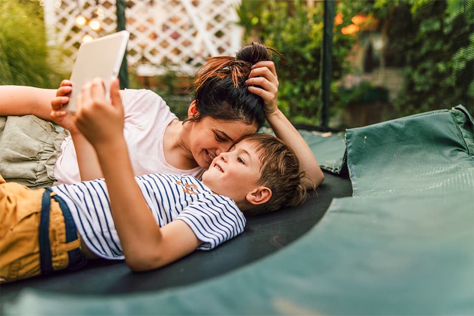 Eine Person und ein Kind liegen gemeinsam auf einem grünen Trampolin und schauen lächelnd auf ein Tablet im Garten.