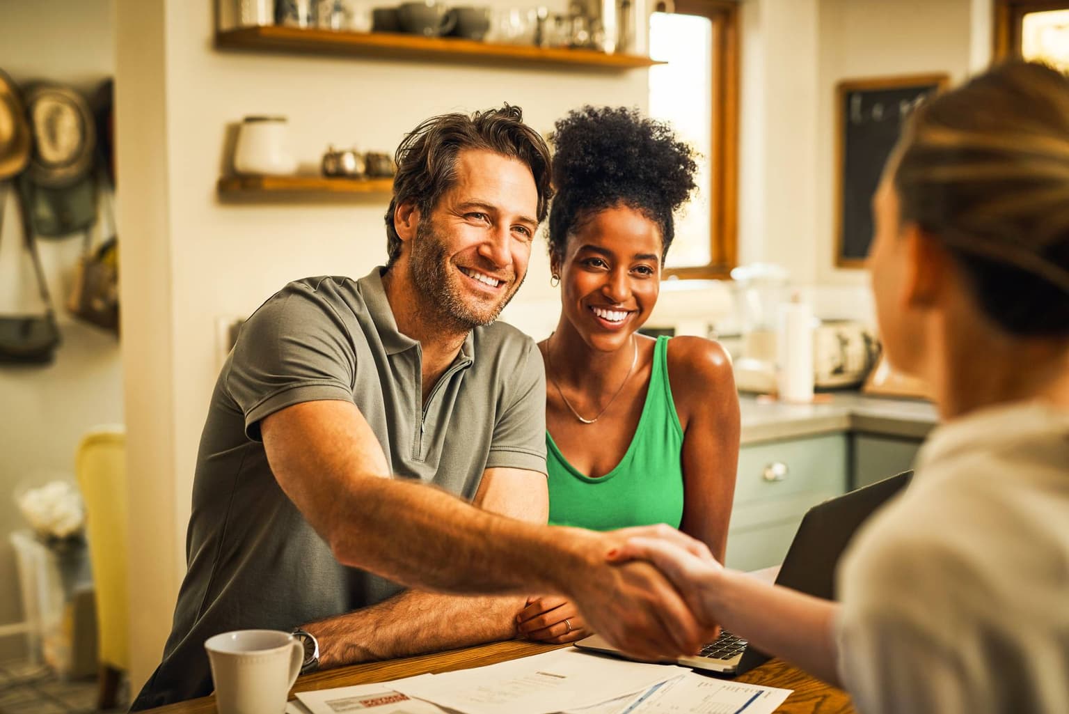Lächelndes Paar in gemütlicher Küche im Gespräch mit einer Person, Dokumente liegen auf dem Holztisch.