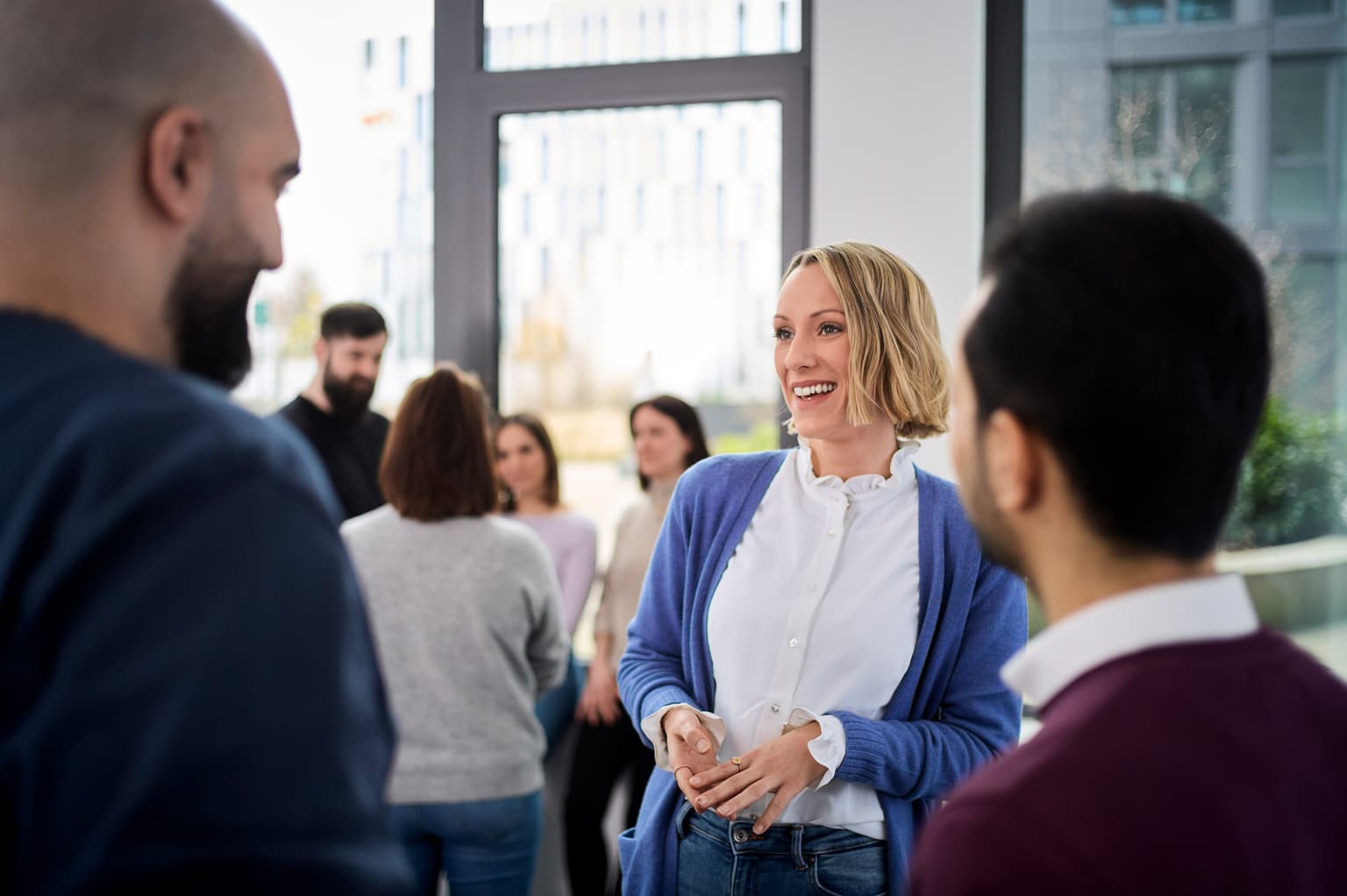 Das Bild zeigt Personen im Gespräch. Der Fokus liegt auf einer Mitarbeiterin, die sich mit zwei Mitarbeitern unterhält. Im Hintergrund ist eine weitere Personengruppe zu sehen, die im Gespräch ist.