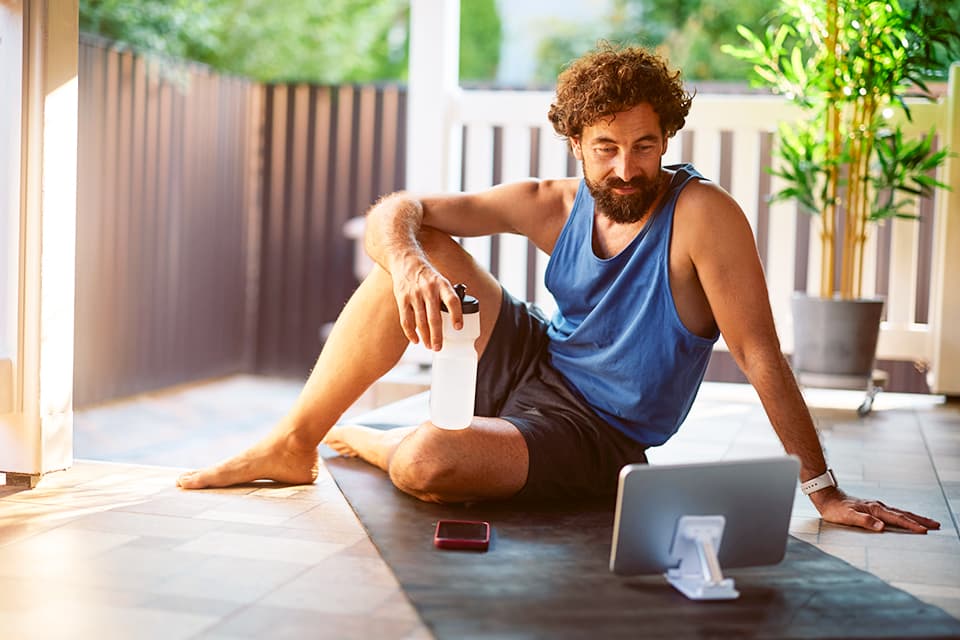 Person in blauem Tanktop sitzt auf Yogamatte mit Wasserflasche, Laptop und Smartphone auf sonniger Terrasse mit Pflanzen.