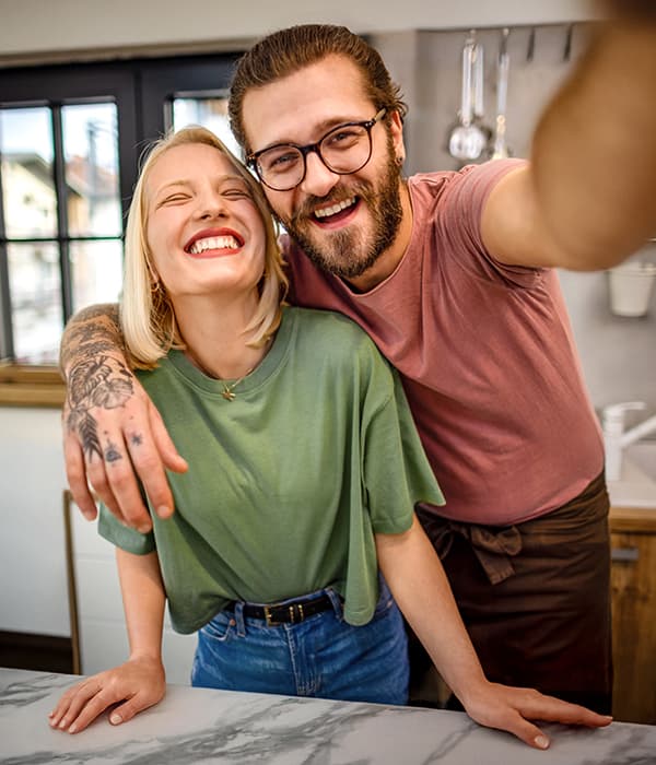Lächelndes Paar macht Selfie in moderner Küche, Person mit Brille trägt rosa Shirt, andere in grünem T-Shirt.