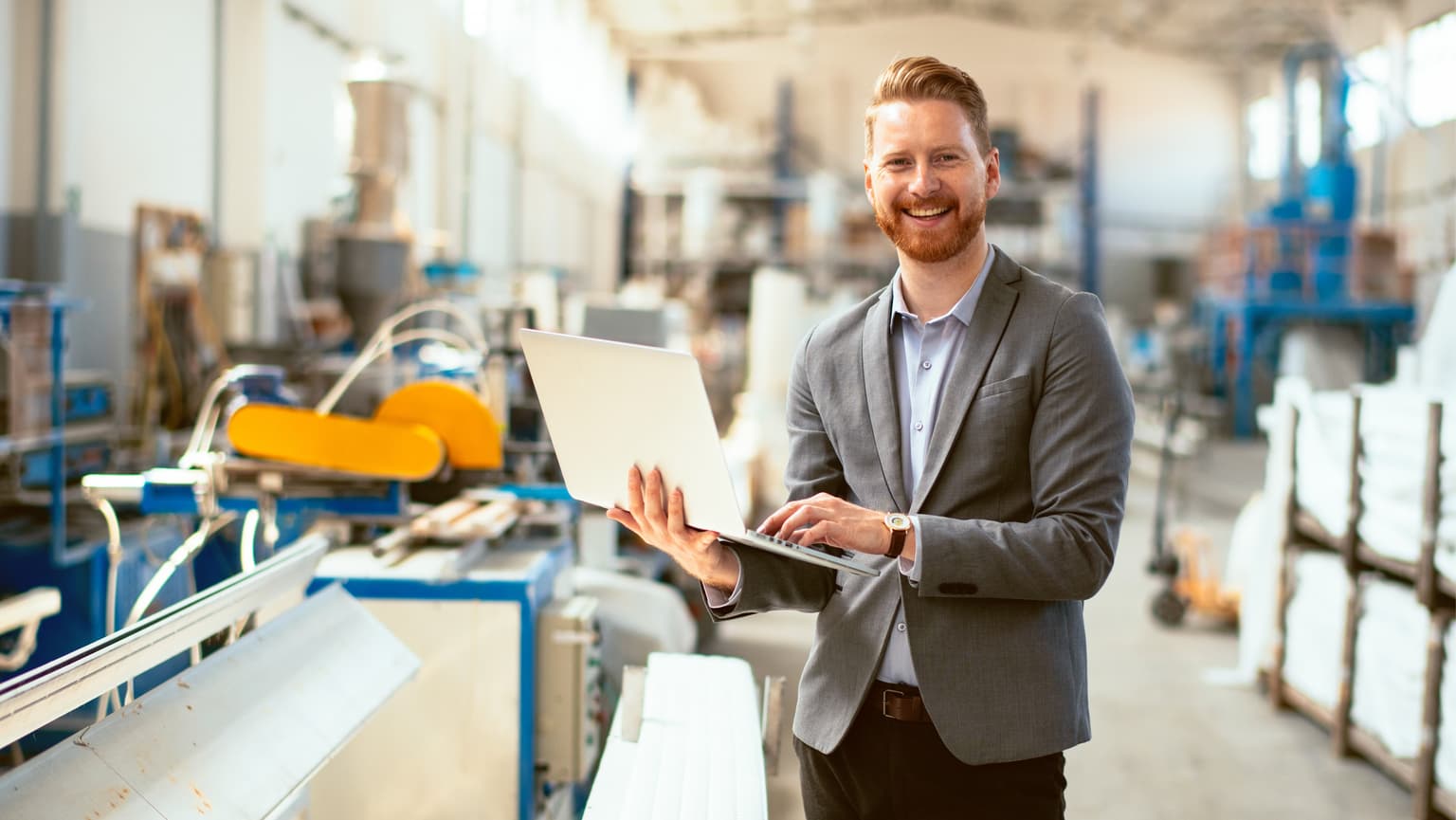Eine Person im Anzug arbeitet an einem Laptop in einer industriellen Umgebung. Im Hintergrund befinden sich verschiedene Maschinen und Anlagen, was auf eine Fabrik oder Produktionsstätte hinweist.