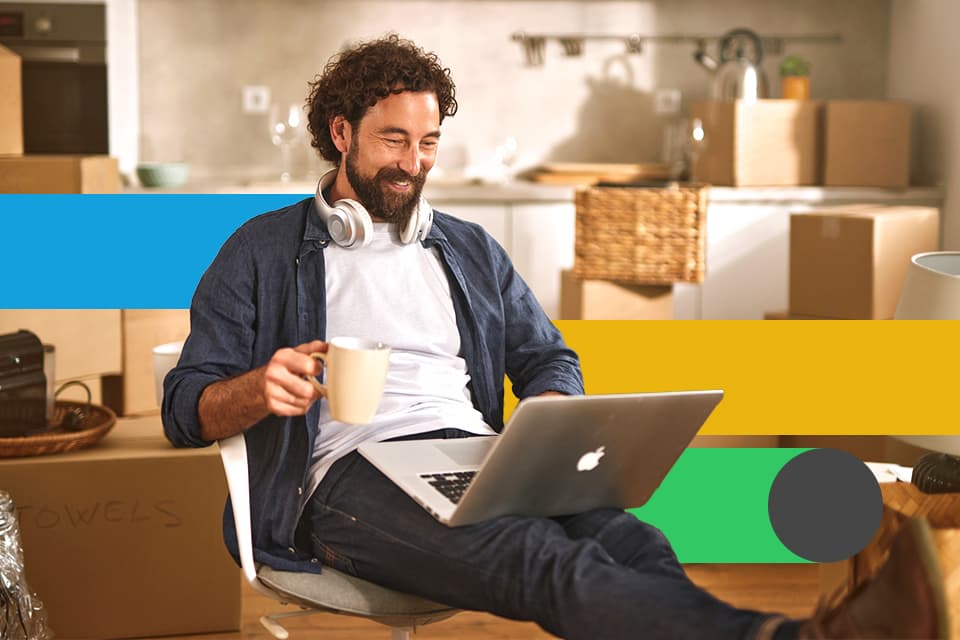 Mann sitzt lächelnd mit Laptop und Kaffeetasse inmitten von Umzugskartons, Kopfhörer um den Hals.