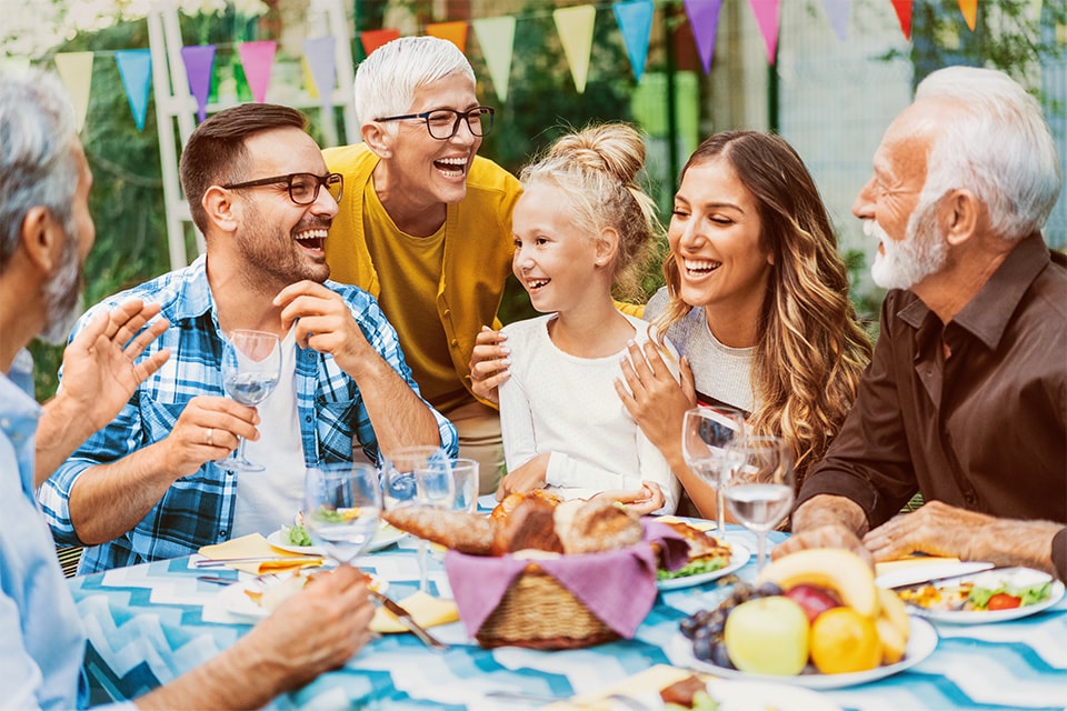 Fröhliche Mehrgenerationenfamilie lacht beim gemeinsamen Essen im Freien, mit bunten Wimpeln im Hintergrund.