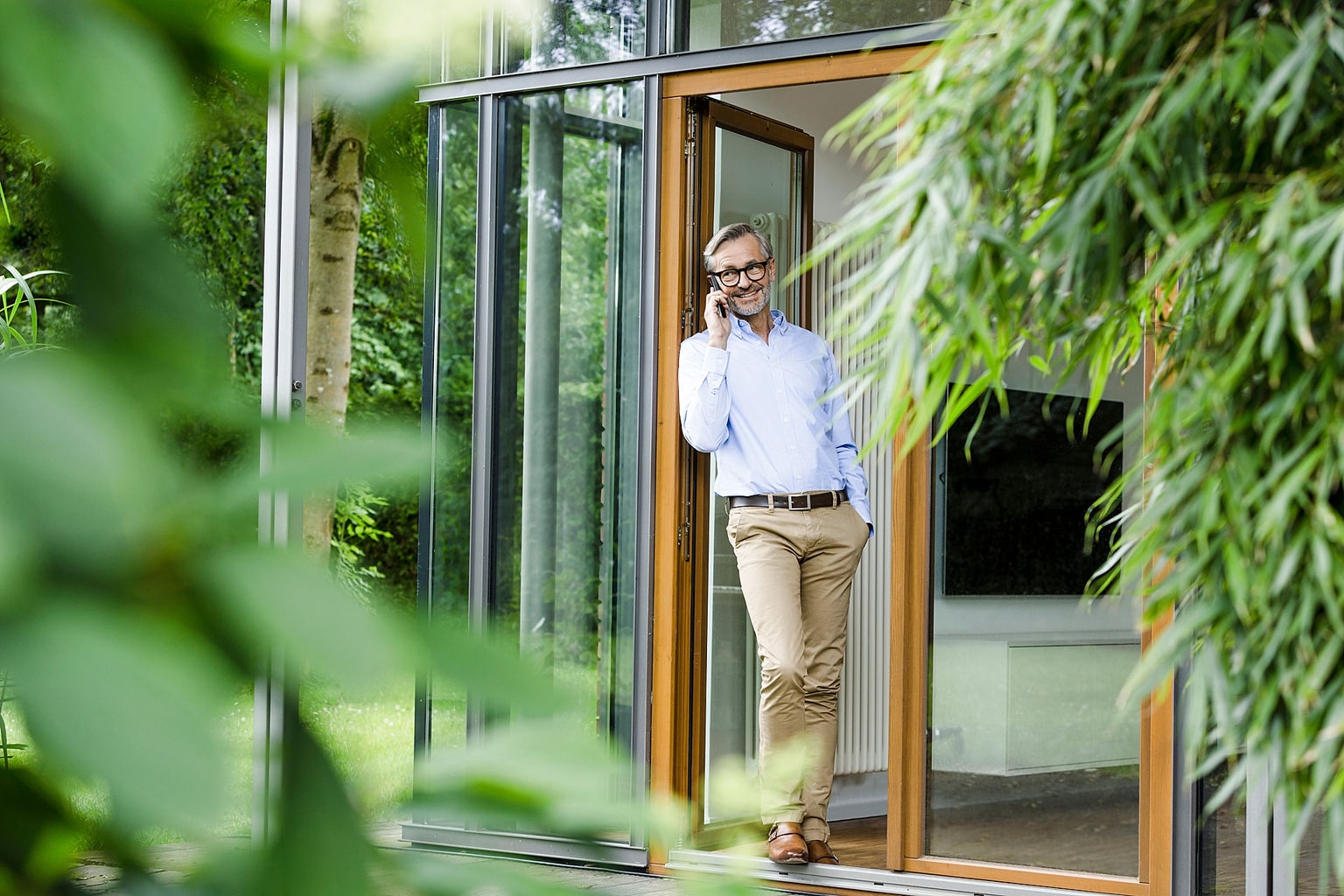 Mann mittleren Alters mit Brille telefoniert lächelnd in einer Glastür eines modernen Hauses, umgeben von grünen Pflanzen.