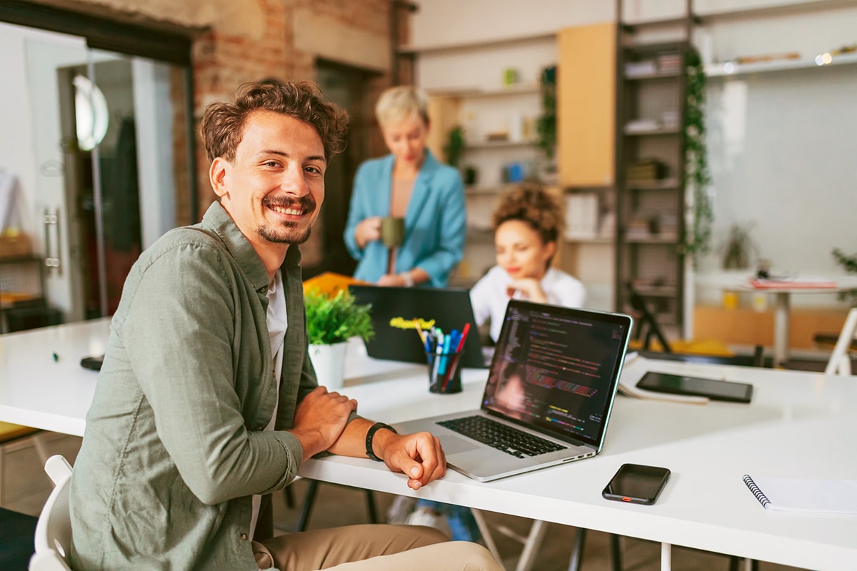 Drei Personen in einem Büro: Eine sitzt an einem Schreibtisch mit einem Laptop, auf dem Code angezeigt wird. Eine weitere Person steht dahinter, eine dritte sitzt weiter im Hintergrund. Im Raum befinden sich Regale mit verschiedenen Gegenständen und Pflanzen sowie weitere Büroartikel auf dem Schreibtisch.