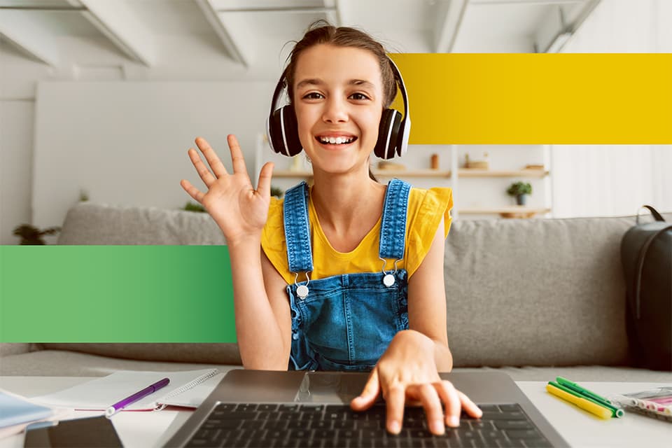 Lächelndes Mädchen mit Kopfhörern und Jeans-Latzhose winkt vor einem Laptop beim Online-Lernen zu Hause.