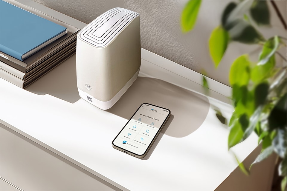 Weißer Router auf Regal neben Smartphone mit App und grüner Pflanze, blaues Buch im Hintergrund.