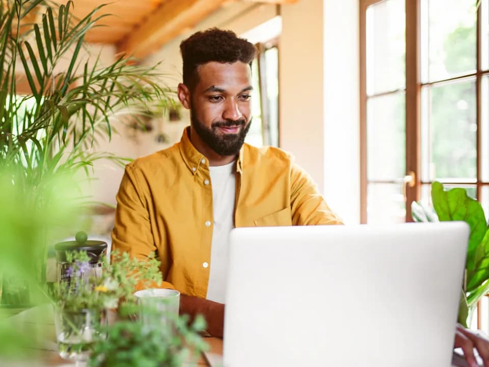 Person in gelbem Hemd arbeitet lächelnd am Laptop in hellem Raum mit Pflanzen und großen Fenstern.