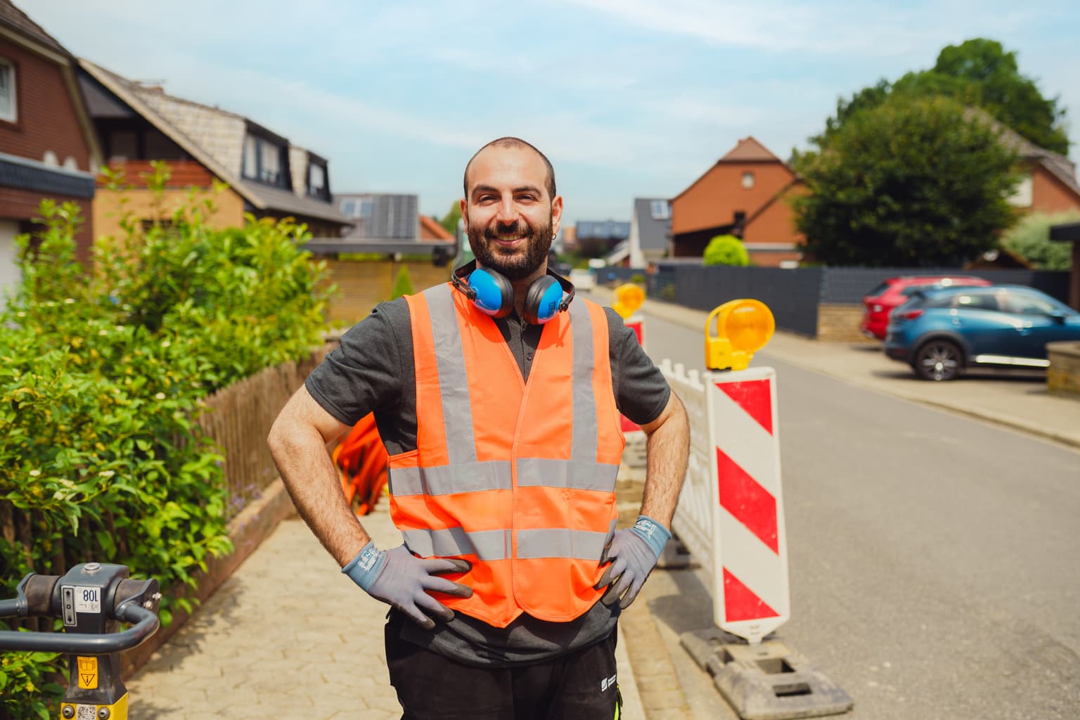 Ein Mann trägt eine orangefarbene Warnweste, blaue Handschuhe und Gehörschutz um den Hals. Er steht mit den Händen in der Hüfte auf einer Wohnstraße. Hinter ihm befinden sich rot-weiß gestreifte Absperrungen mit orangefarbenen Warnleuchten. Im Hintergrund sind Häuser und parkende Autos zu sehen.