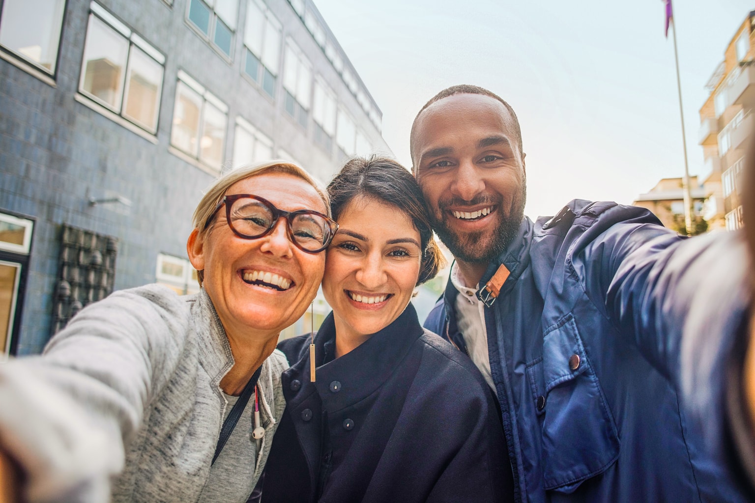 Drei lächelnde Personen machen ein Selfie in einer Stadtstraße. Zwei Frauen und ein Mann in Freizeitkleidung, Gebäude im Hintergrund.