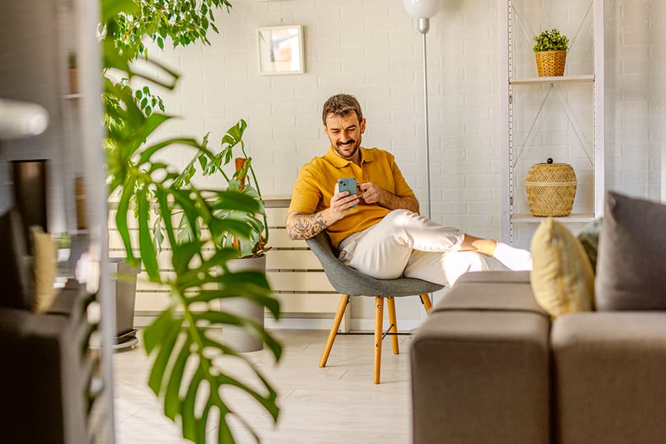 Person in gelbem Hemd sitzt im modernen Wohnzimmer mit Pflanzen und nutzt ein Smartphone.