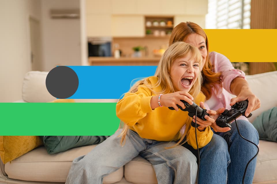 Mädchen und Frau spielen begeistert Videospiele auf dem Sofa mit schwarzen Controllern in bunter Wohnzimmeratmosphäre.