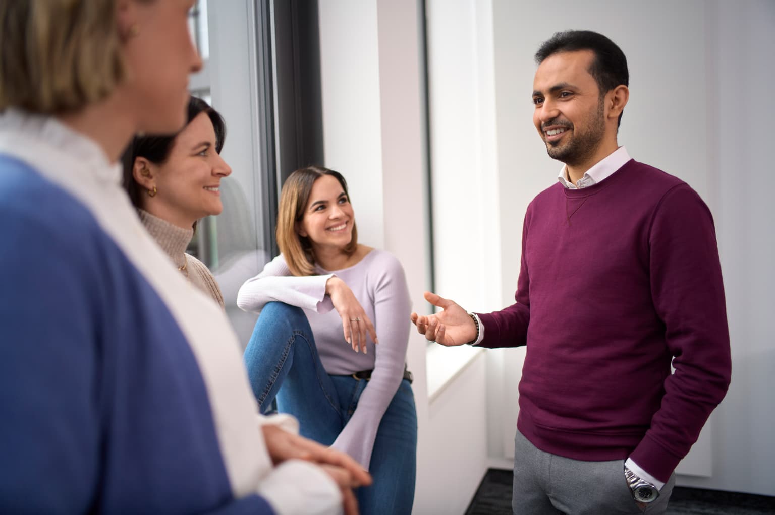 Diversität bei deutsche Glasfaser Mitarbeiter. Vier Personen im Gespräch in einem Büro oder Besprechungsraum. Eine Person steht und gestikuliert mit der Hand, während die anderen aufmerksam zuhören.