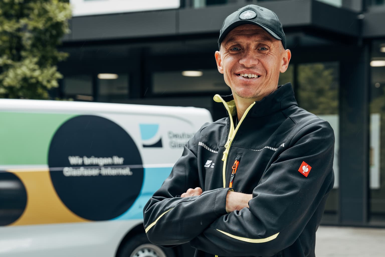 Das Bild zeigt einen Mitarbeitenden von Deutsche Glasfaser. Er ist im Bereich der Glasfasermontage tätig und trägt Arbeitskleidung von Deutsche Glasfaser. Er steht vor dem Bürogebäude in Düsseldorf. Zudem ist auch noch ein Montagefahrzeug von Deutsche Glasfaser im Hintergrund zu erkennen.