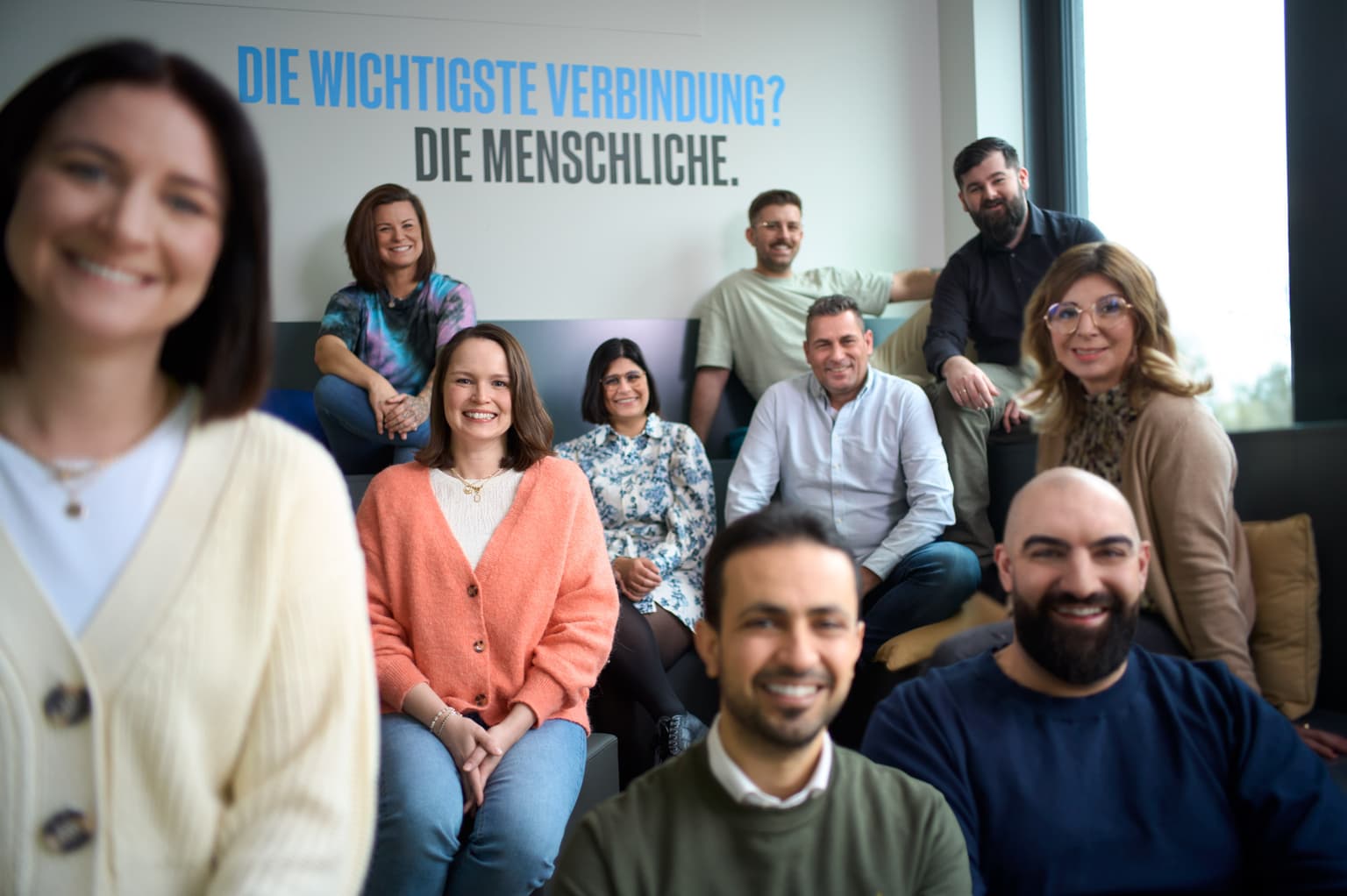Das Bild zeigt eine Gruppe von Mitarbeitenden, die auf einer Tribüne sitzen und fröhlich in die Kamera lächeln. Auf der Wand im Hintergrund steht "Die wichtigste Verbindung? Die Menschliche."
