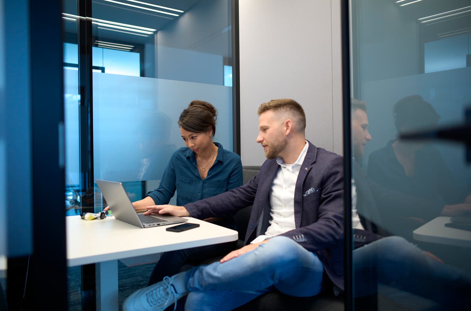 Das Bild zeigt zwei Mitarbeitende an einem Arbeitsplatz. Beide sind in einem Gespräch und schauen sich gemeinsam etwas auf dem Bildschirm an. Im Hintergrund sind weitere Personen an Arbeitsplätzen zu sehen.