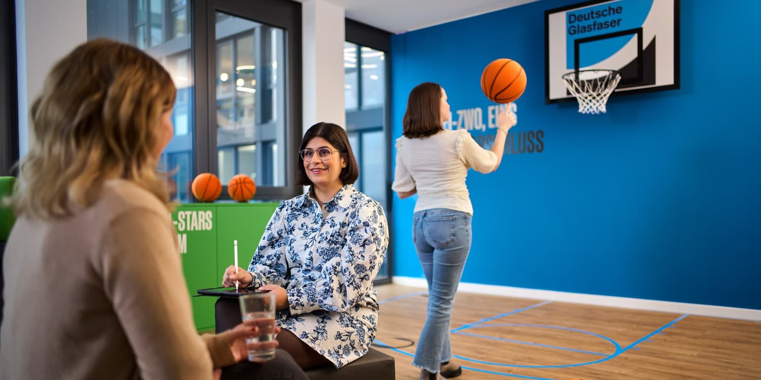 Das Bild zeigt 3 Personen in einem Raum, der über einen Basketballkorb verfügt. Eine Person hat einen Basketball in der Hand. Sie ist im Hintergrund zu sehen. Die anderen beiden Personen sitzen im Vordergrund. Eine Person ist mit dem Rücken zur Kamera gedreht. Die andere Person hält ein Tablet und einen Stift in der Hand.