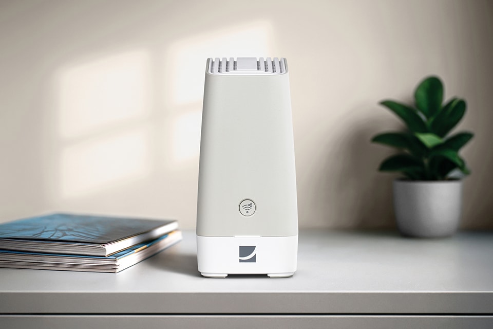 Weißer WLAN-Router auf einem Tisch neben Zeitschriften und einer kleinen Zimmerpflanze im Hintergrund.