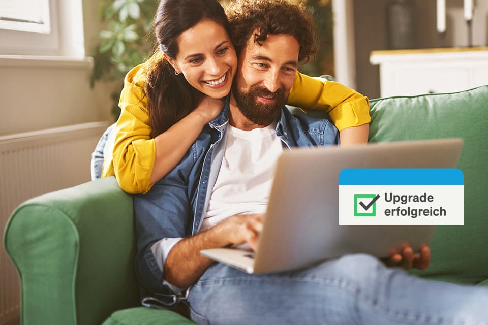 Ein Paar sitzt lächelnd auf einem grünen Sofa mit einem Laptop. Eine Meldung zeigt "Upgrade erfolgreich" mit grünem Häkchen.