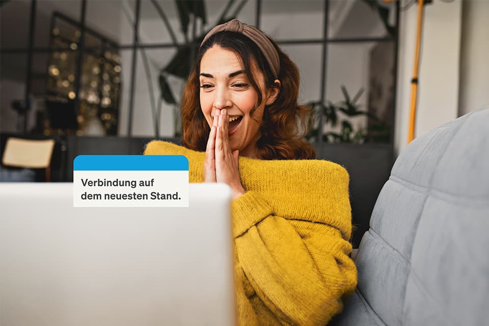 Begeisterte Frau im gelben Pullover sitzt auf Sofa vor Laptop. Text: „Verbindung auf dem neuesten Stand."