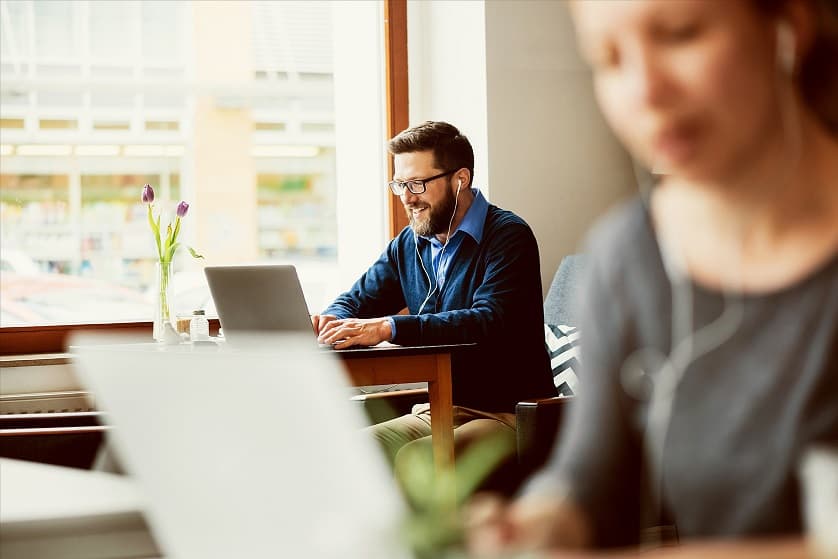 Szene in einem Café oder ähnlichen Umfeld, in dem zwei Personen an Laptops arbeiten. Eine Person sitzt am Fenster an einem Tisch mit einem Laptop und trägt ein blaues Shirt. Die andere Person befindet sich im Vordergrund, ebenfalls mit einem Laptop. Auf dem Tisch am Fenster steht ein Blumenstrauß, im Hintergrund sind unscharfe Elemente einer städtischen Umgebung zu erkennen.