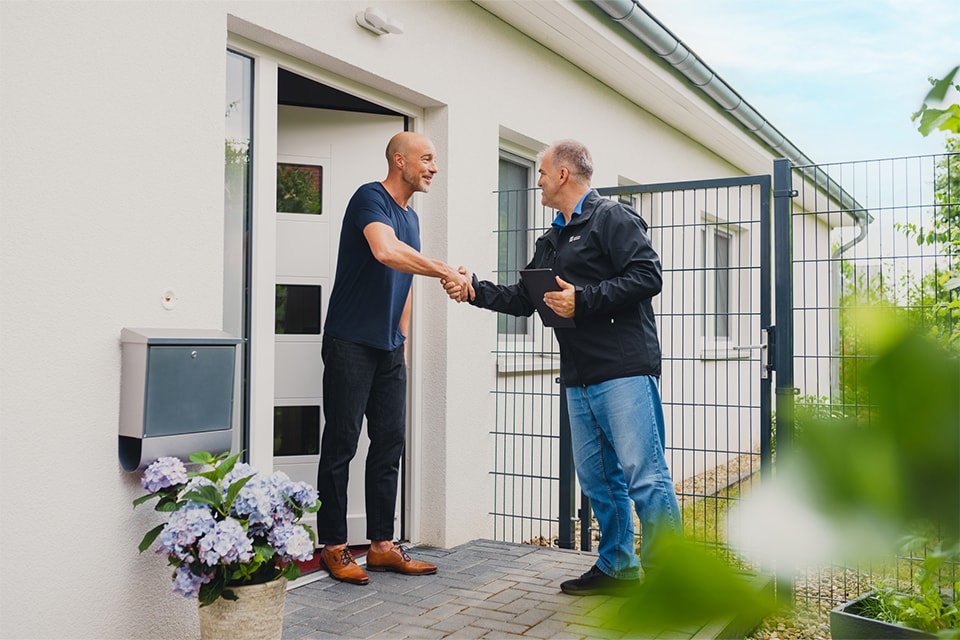 Zwei Personen geben sich die Hand am Eingang eines Hauses. Eine Person steht im Türrahmen, die andere draußen auf einem gepflasterten Bereich. Neben der Tür steht ein Blumentopf mit Blumen, im Hintergrund ist ein Zaun zu sehen.