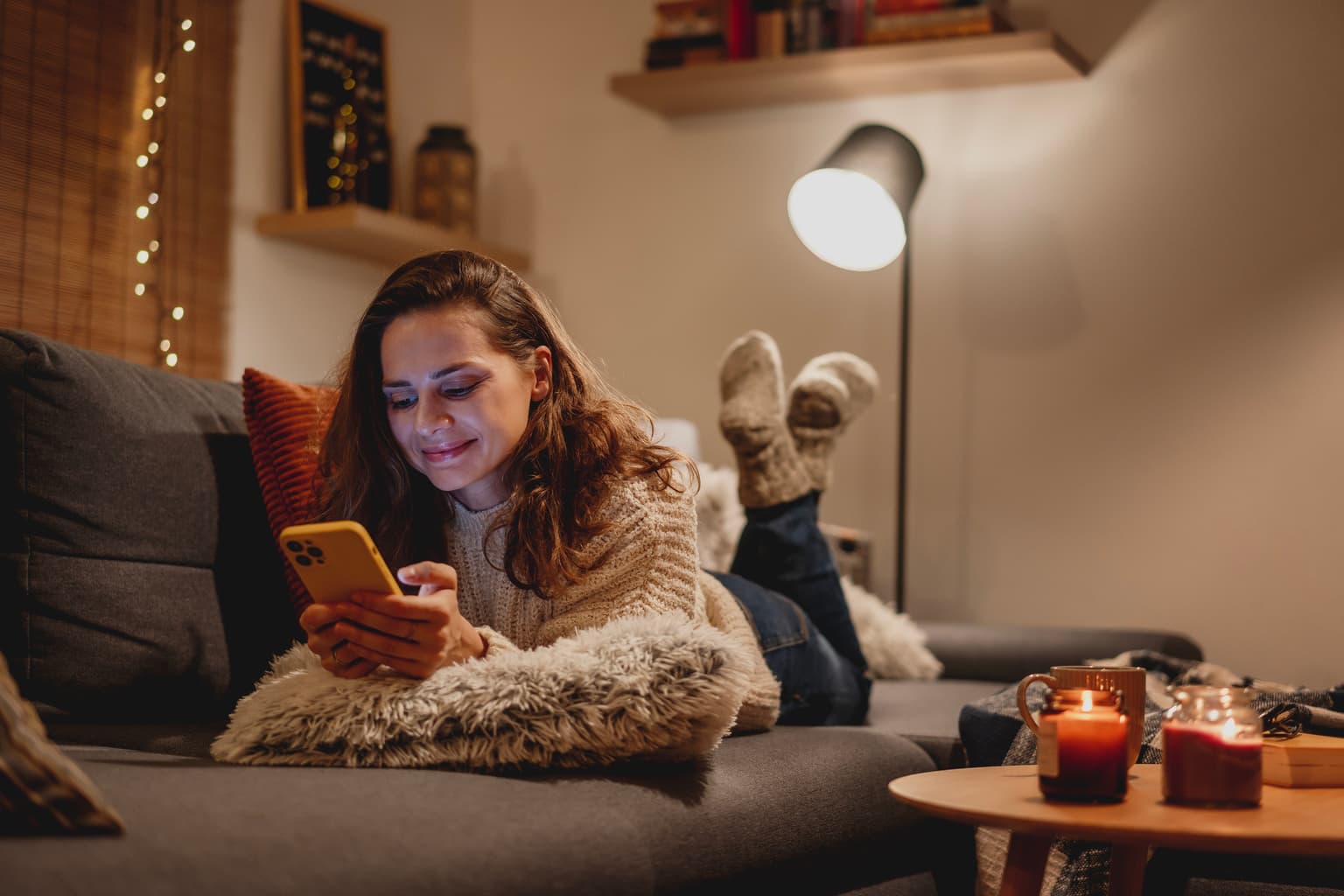 Frau liegt auf einem Sofa, trägt weißen Pullover und Socken, hält ein gelbes Smartphone. Beine hochgelegt, im Vordergrund ein kleiner Tisch mit brennenden Kerzen, im Hintergrund eine eingeschaltete Stehlampe. Gemütliche Atmosphäre mit warmem Licht, Kissen und dekorativen Elementen wie Regalen.