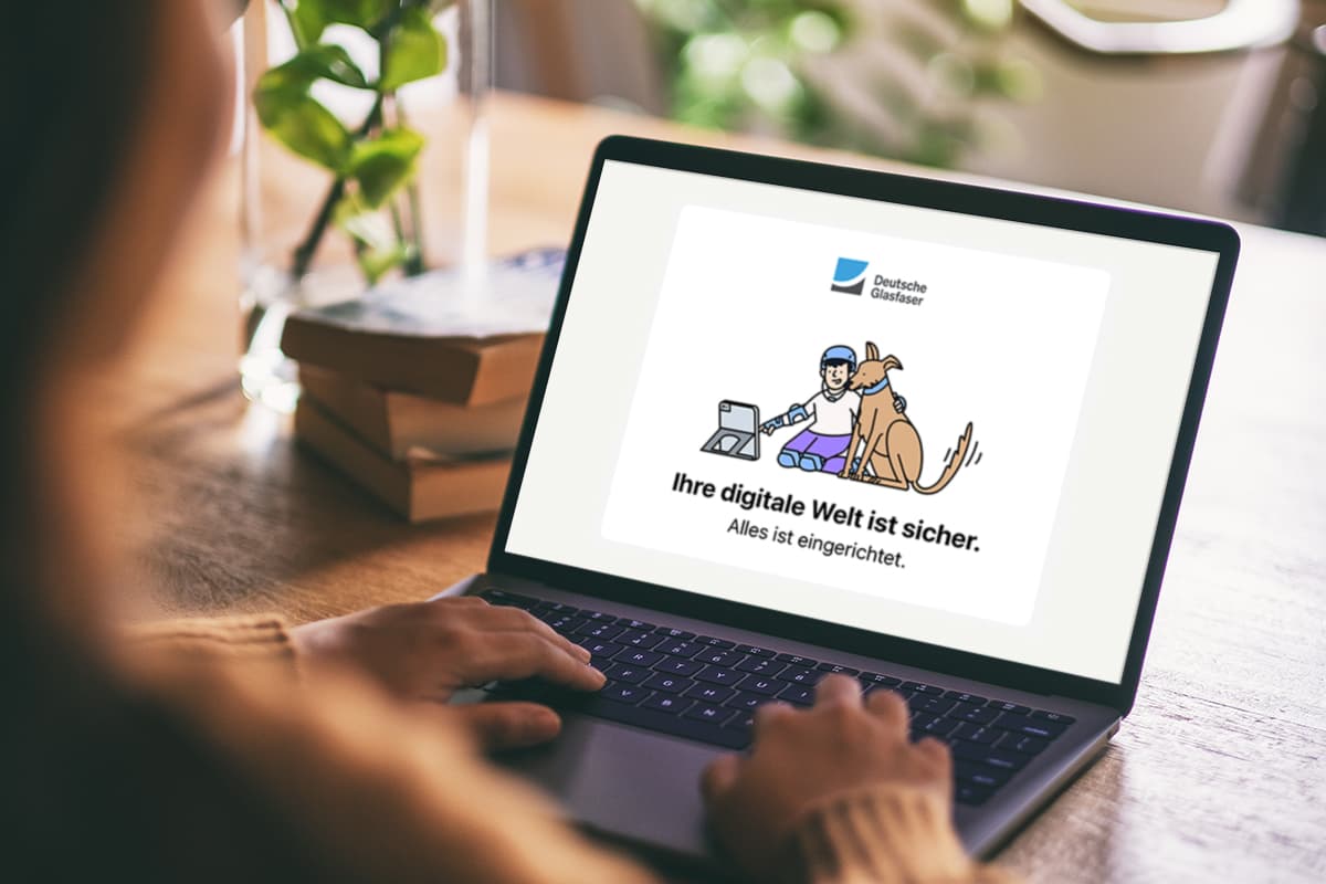 Eine Person arbeitet an einem Laptop. Auf dem Bildschirm ist eine Illustration zu sehen: Ein Junge und ein Hund sitzen nebeneinander mit einem Tablet. Darüber steht der Text ‚Deutsche Glasfaser‘. Darunter steht: ‚Ihre digitale Welt ist sicher. Alles ist eingerichtet.‘