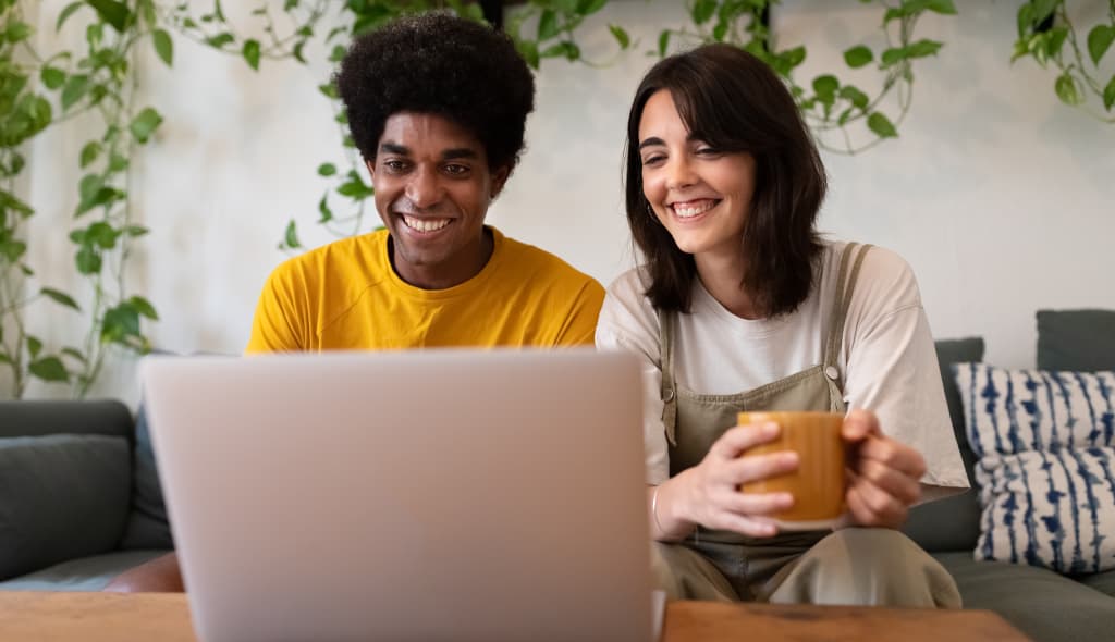 Beratung Zuhause - Paar sitzt auf der Couch und vereinbart einen Termin online auf dem Laptop