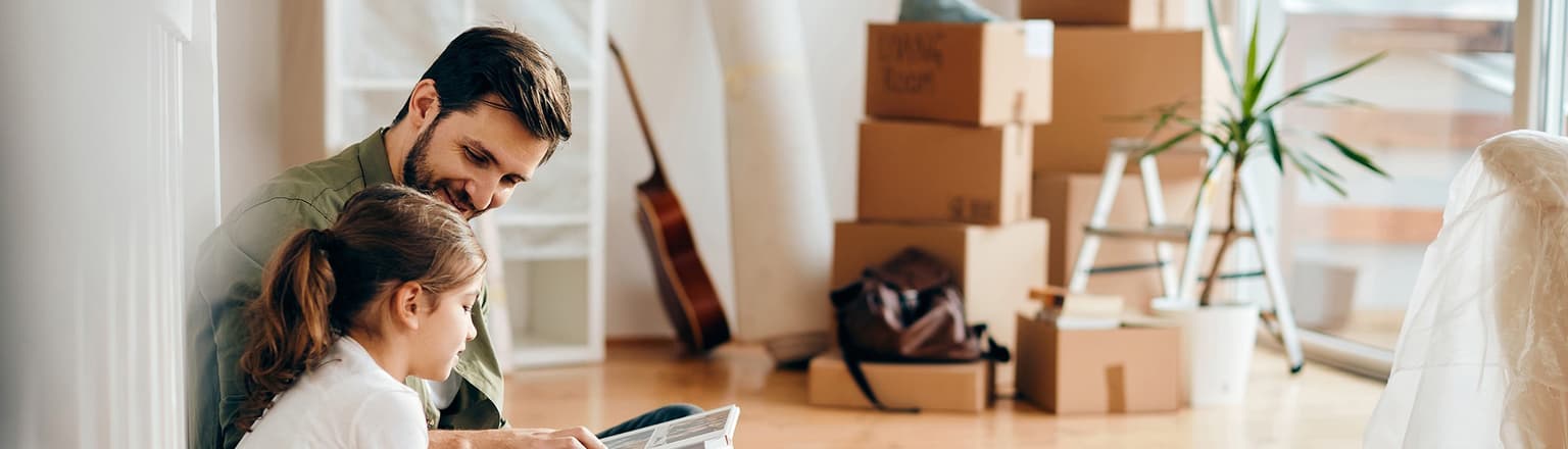 Mann sitzt mit Tochter auf dem Boden und blättert lachend durch ein Fotobuch - Umzugskisten im Hintergrund