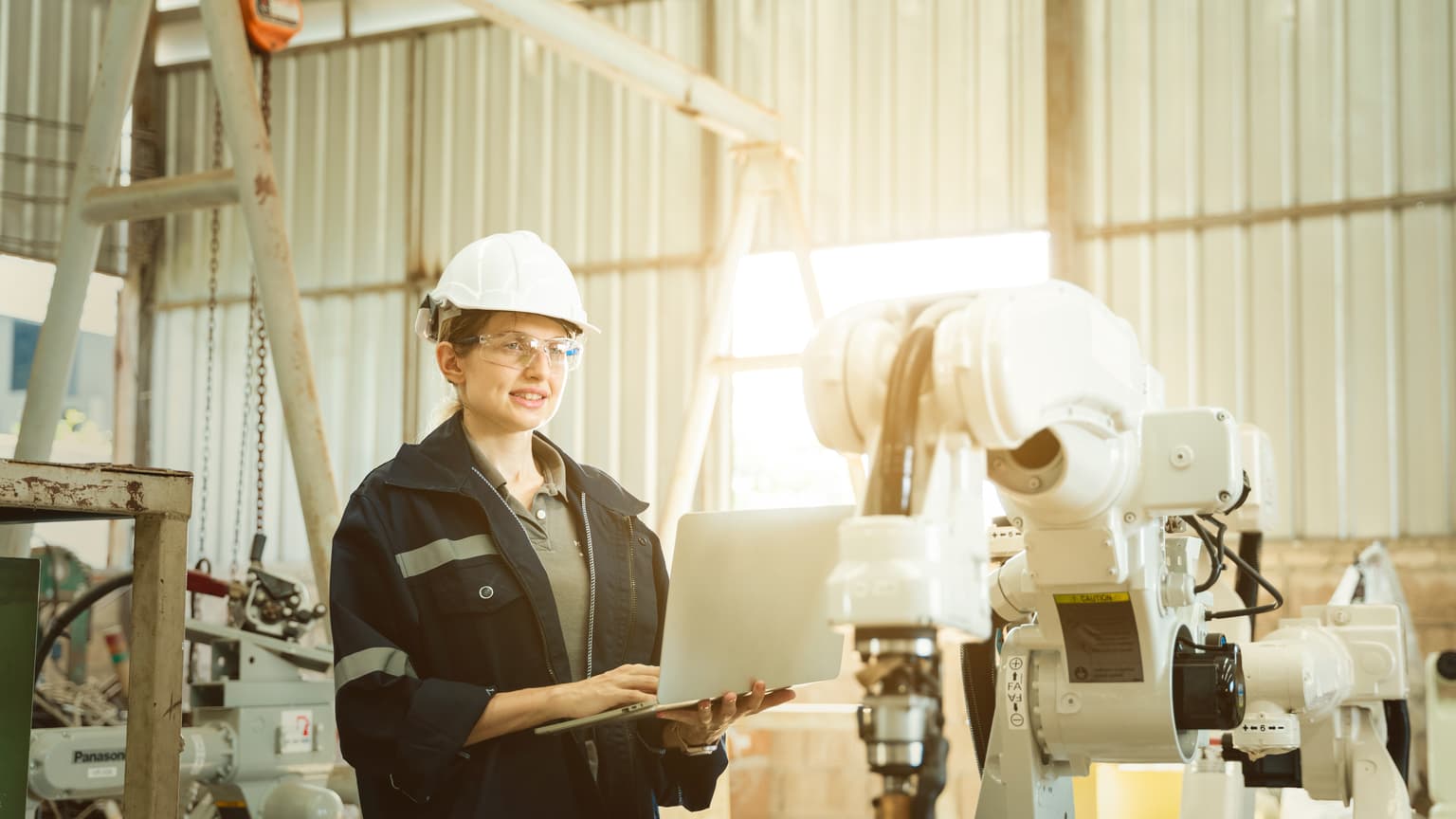 Geschäftsprozesse optimieren & digitalisieren - Frau mit Sicherheitsbrille und Helm arbeitet in einer Lagerhalle an einem Roboterarm
