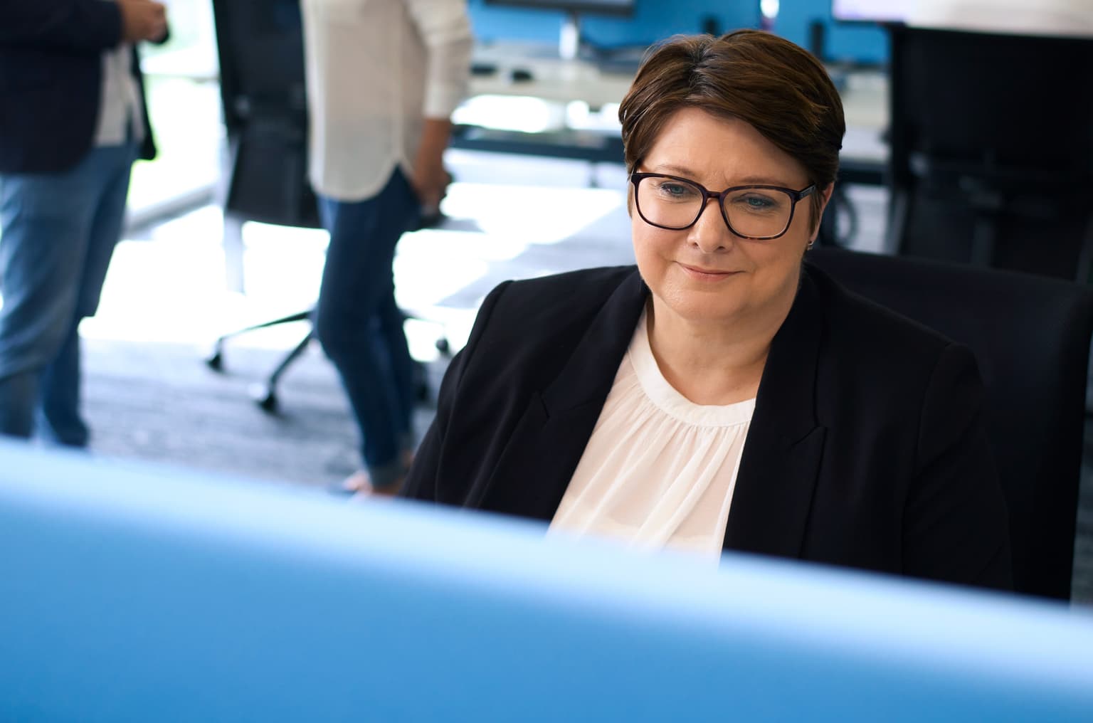 Das Bild zeigt eine Mitarbeiterin von Deutsche Glasfaser, die am Schreibtisch sitzt und arbeitet. Das Bild ist im Büro aufgenommen. Im Hintergrund sind andere Mitarbeiterinnen und Mitarbeiter zu sehen, die ebenfalls arbeiten oder sich unterhalten. Der Hintergrund ist unscharf.