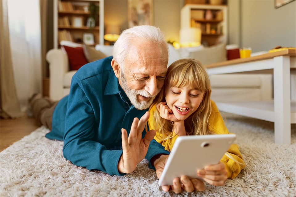 Älterer Mann und junges Mädchen liegen lachend auf dem Boden und winken gemeinsam in ein Tablet bei einem Videoanruf.