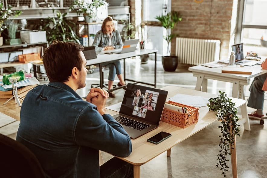 Modernes Büro mit mehreren Personen, die an Schreibtischen arbeiten. Im Vordergrund nimmt eine Person an einer Videokonferenz über einen Laptop teil. Im Hintergrund arbeiten weitere Personen an ihren Arbeitsplätzen. Das Büro ist offen gestaltet, mit Pflanzen und natürlichem Licht durch große Fenster.