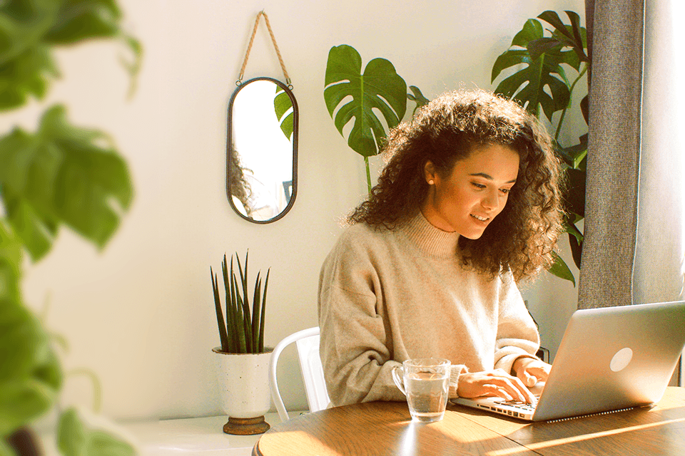 Frau mit lockigem Haar arbeitet am Laptop in hellem Raum mit Zimmerpflanzen, Spiegel und gemütlicher Atmosphäre.