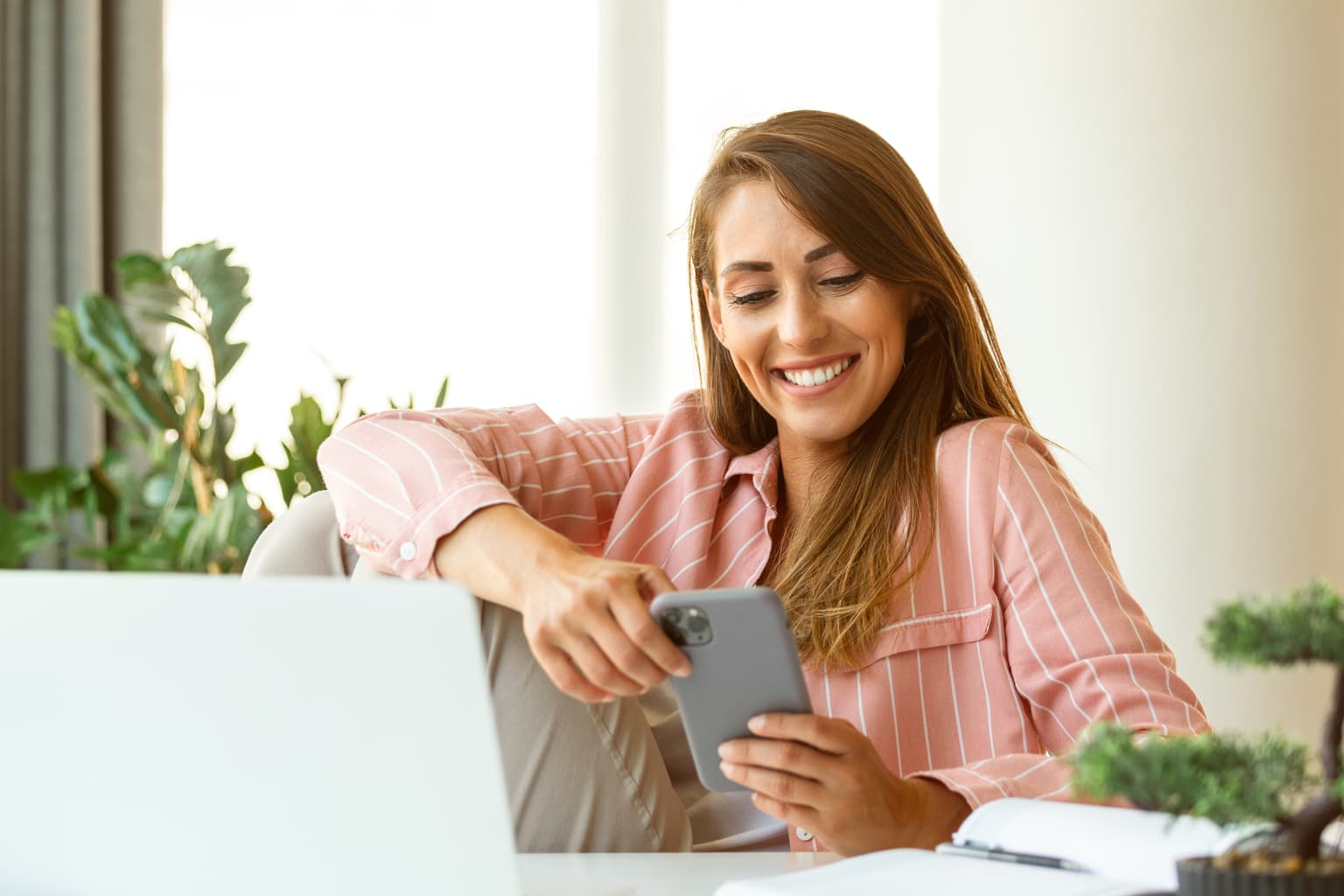 Ihre Rolle in der Bauphase - Frau im pink-gestreiften Blazer sitzt am Tisch und schat lächelnd auf das Handy