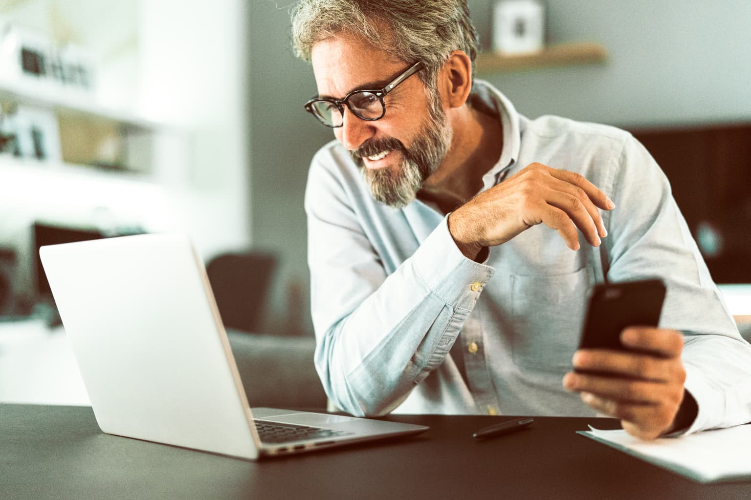 Älterer Mann hält Handy in der Hand und schaut auf den Laptop
