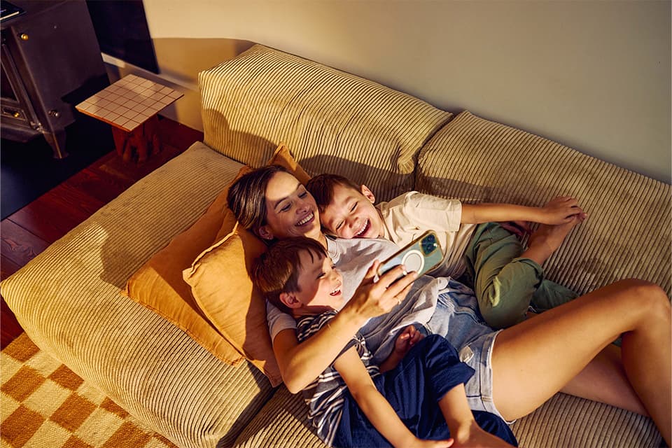 Mutter liegt lachend mit zwei Söhnen auf einem beigen Sofa und schaut gemeinsam auf ein Smartphone.