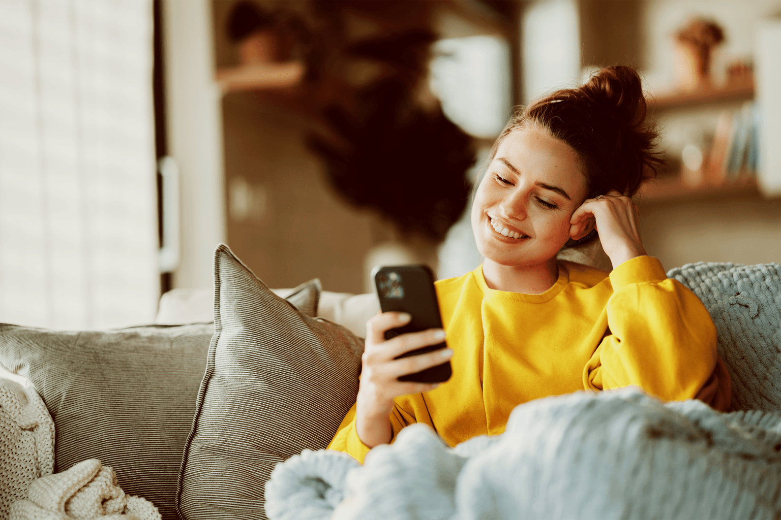 Person in gelbem Pullover lächelt beim Betrachten des Smartphones auf gemütlichem Sofa mit grauen Kissen.