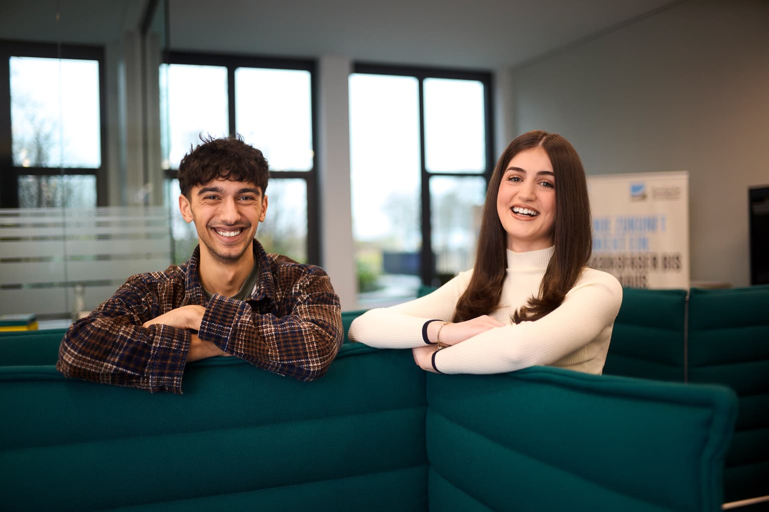 Das Bild zeigt zwei Mitarbeitende, die sich im Workshopraum befinden. Im unteren Drittel des Bildes ist eine Couch mit hohen Lehnen zu sehen. Die Mitarbeitenden (Frau und Mann) stehen hinter der Couch und stützen sich mit den Armen auf dieser ab. Beide lächeln in die Kamera. Im Hintergrund sind Fenster und ein Aufsteller verschwommen zu sehen.