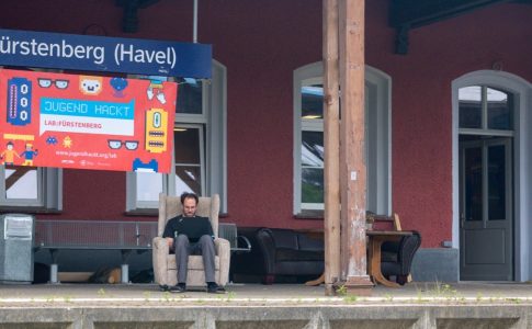 Ein Mann sitzt in einem Sessel auf dem Bahnsteig, über ihm das Schild „Fürstenberg (Havel)“ und eine Werbetafel für die Veranstaltung „jugend hackt“