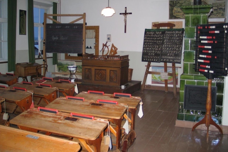 Ein historischer Dorfschul-Klassenraum mit Holzbänken, einem Rechenbrett und einem Kachelofen.