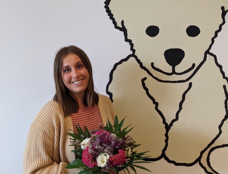 Eine junge Frau steht mit einem Blumenstrauß neben einer Zeichnung eines Bären